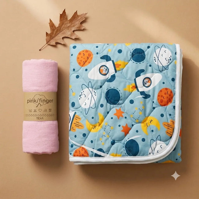 A folded light blue quilted blanket with a playful space print featuring rockets, astronauts, planets, stars, and moons, displayed next to a rolled pink baby towel wrapped in a pink paper band labeled 'pink/finger'. The items are styled on a warm beige background with a decorative dried leaf.